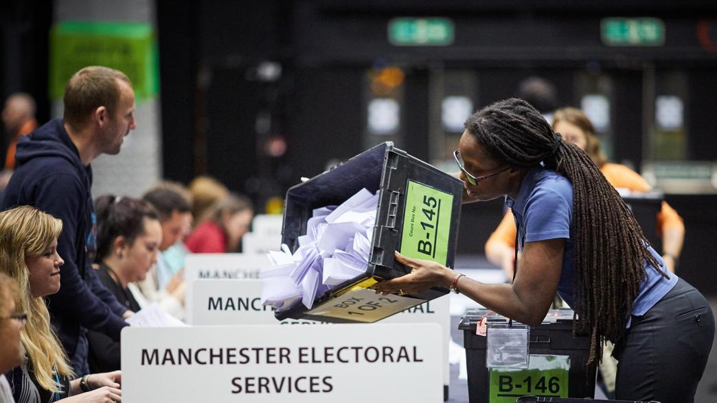 A woman at an election result vote count tipping over a box of ballot papers onto the table.
