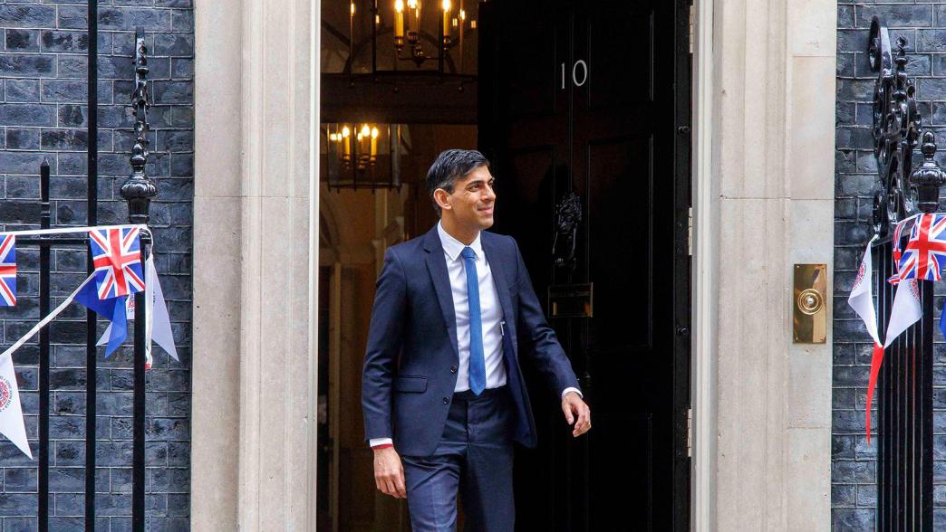 Rishi Sunak walking out the door of No.10 Downing Street. There is blue, red, white and Union Jack bunting outside the building for King Charles III coronation.
