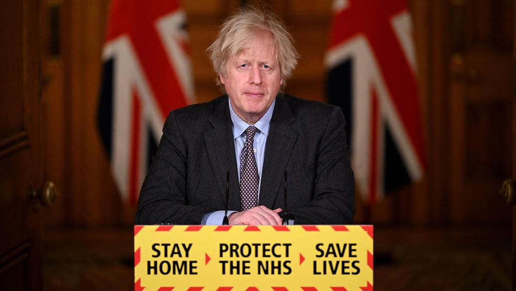 Boris Johnson during a coronavirus press briefing in 2020. He is stood behind a podium which has the messaging: Stay Home. Protect the NHS. Save Lives