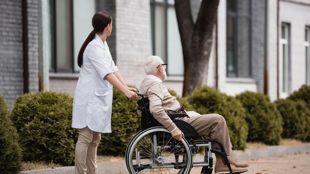 A female social worker walking outside with a man in a wheelchair. Both are faced away from the camera.
