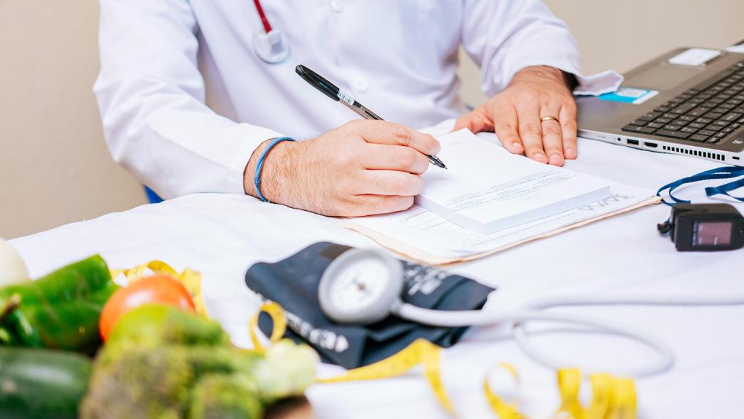 A close up of a medical expert taking notes. On the desk is a plate with green vegetables, a blood pressure machine and a laptop.