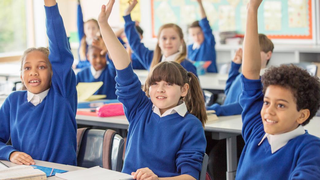 Primary school children wearing blue school uniforms raising hands in classroom.