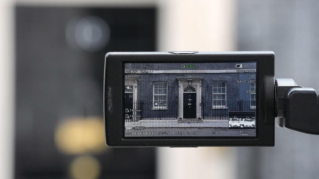A screen of a TV camera pointing at the 10 Downing Street door is standing in the street in London