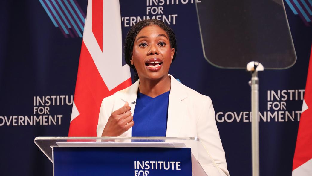 Kemi Badenoch. She is wearing a blue dress and a white blazer. She is standing on stage at the Institute for Government.