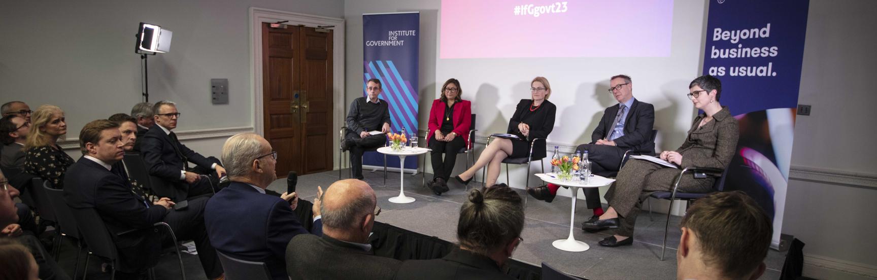 Panellists on stage at an event at the IfG Government 2023 conference, including Ayesha Hazarika, Paul Johnson and Chloe Smith MP while a member of the audience asks a question.