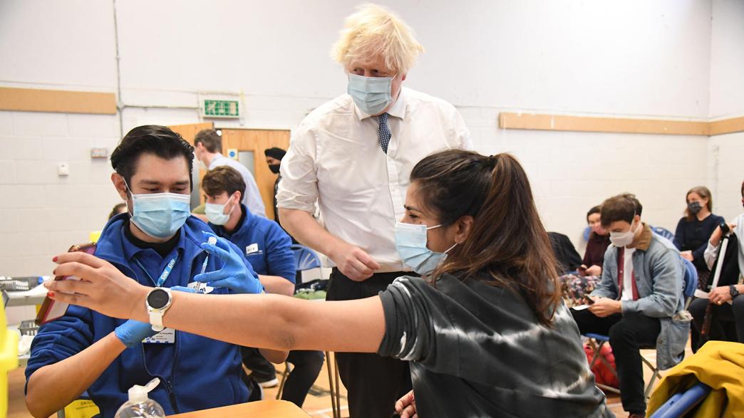 Boris Johnson wearing a face mask on a visit to a Covid vaccine centre. A women has her sleeve rolled up as a medical assistant prepares to give her the vaccine.